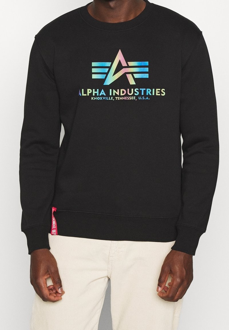 Felpa nera con logo "Alpha Industries" multicolore, polsini e orlo a costine, e un'etichetta rossa sul lato.