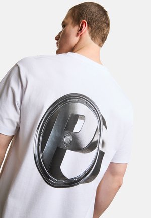 Młody mężczyzna zwrócony tyłem, ubrany w białą koszulkę z dużym metalicznym srebrnym okrągłym emblematem z wyraźną literą "P" na plecach.