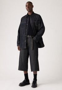 Camisa de mezclilla oscura de corte oversize con costuras visibles, combinada con pantalones de mezclilla cortos. Llevada con una camiseta negra y zapatos negros.