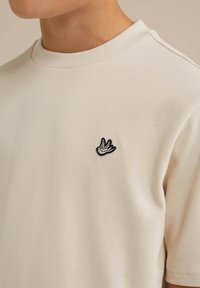 Beige pamut póló kerek nyakkivágással és rövid ujjakkal. A mellkason egy kicsi, fekete-fehér hímzett madármintázat található.
