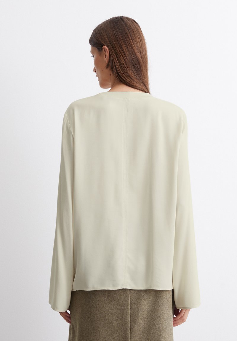 Langärmlige, cremefarbene Bluse mit glatter Textur, geradem Saum und minimalistischem Design. Der Rücken weist ein zentrales Nahtdetail auf.