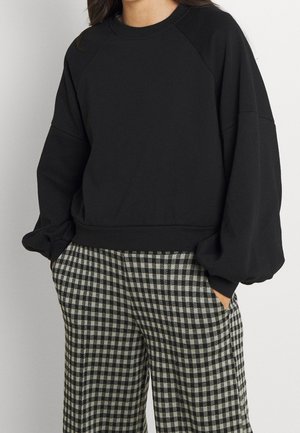 Kvinde iført en sort sweatshirt med lange ærmer og puffede skuldre og sort-hvide ternede bukser, hænderne i lommerne.
