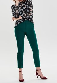 Zielone dopasowane spodnie o gładkiej fakturze, zestawione z czarną bluzką w kwiaty. Ubrane burgundowe sandały na wysokim obcasie.