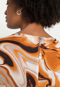 Večbarvna bluzica s spiralo podobnim vzorcem v oranžni, črni in kremni barvi; vključuje majhen moder kvadraten poudarek na zadnjem zaklepu vratu.