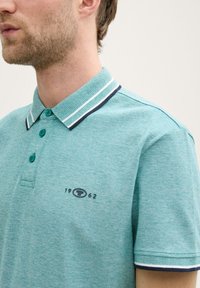 Teal polo skjorta i texturerad tyg med en tvåknappsknäppning, kontrasterande randig krage och broderad logotyp med "1962."
