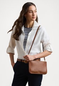 Borsa a tracolla in pelle marrone con una texture liscia, con un logo dorato, portata con una tracolla. Design semplice e rettangolare.