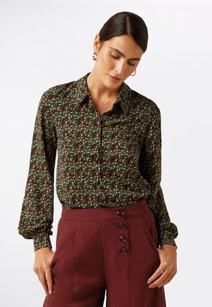 Long-sleeve blouse in dark fabric with a vibrant leaf pattern in green, yellow, and red. Features button closure and a pointed collar.