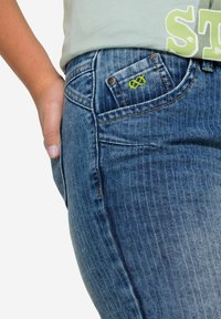 Jeans i medelblå denim med ett subtilt vertikalt stripemönster. Har en liten grön oändlighetssymbol på framfickan och orange sömmar.