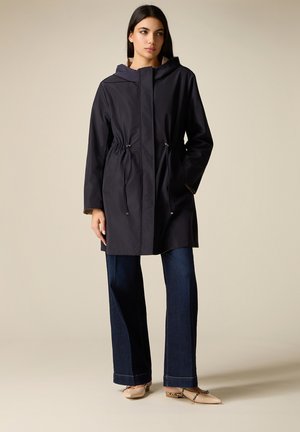 Frau trägt einen schwarzen Kapuzenmantel mit Kordelzug, dunkelblaue Schlagjeans und beige spitze Slipper mit Knöchelriemen, steht vor einem einfarbigen Hintergrund.