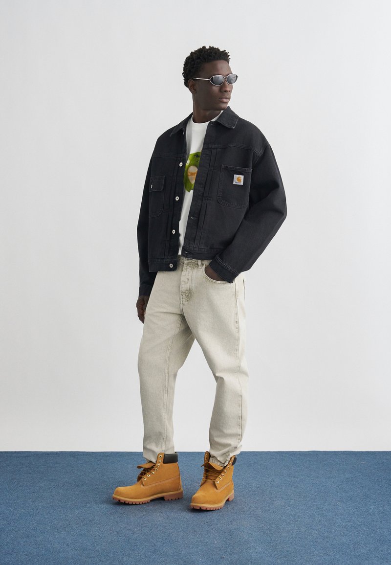 Homme portant une veste noire, un t-shirt blanc avec un motif graphique, un pantalon beige clair, des bottes couleur cuir clair et des lunettes de soleil, debout sur un sol bleu devant un mur gris.