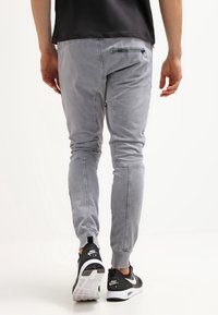 Šedé zúžené tepláky s elastickými manžetami, vybavené zipovou kapsou a hladkou strukturou, spárované s černými teniskami.