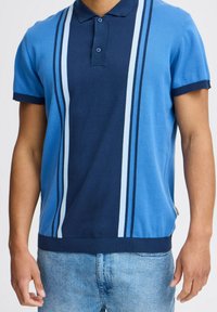 Kék póló, sötétkék és világoskék függőleges csíkokkal, rövid ujjakkal és klasszikus gallérral. Puha kötött anyagból készült.