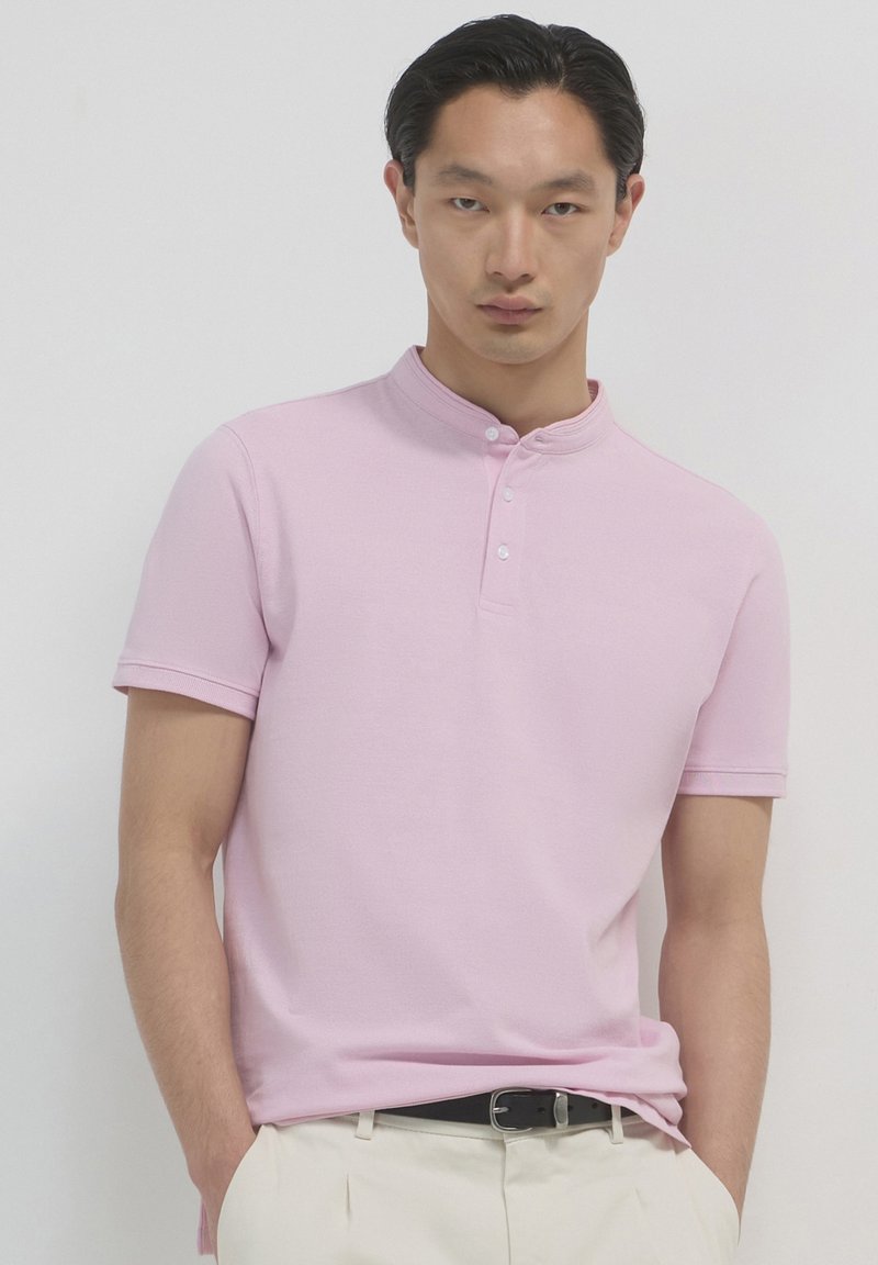 Polo shirt rosa chiaro a maniche corte con colletto alla mandarino e patta con tre bottoni. Tessuto morbido con una texture liscia. Abbinato a pantaloni chiari.