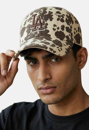 Man wearing beige and brown spotted cap with dark "LA" logo, adjusting the brim, wearing a black shirt, neutral background.