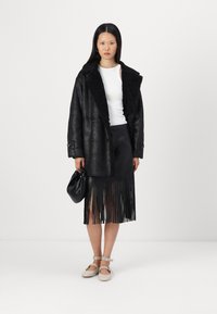 Fekete műbőr kabát, texturált, bolyhos gallérral, fekete fröccsöntött szoknyával és bézs cipővel párosítva. Divatos kézitáska a kezében.