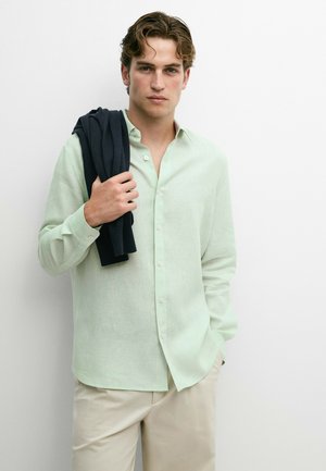 Junger Mann trägt ein hellgrünes Hemd mit Knöpfen und beige Hose, steht und hält einen dunklen Pullover über der Schulter vor weißem Hintergrund.