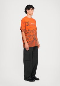 Orange grafisk t-shirt med landskabsprint, korte ærmer og løs pasform, parret med sorte cargo-bukser og sorte sko.
