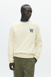 Homme portant un sweat crème avec un logo "W" bleu et un pantalon vert, regardant vers sa gauche avec une main dans la poche.