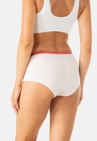 Femme portant des sous-vêtements blancs à motifs avec une bande rose et un soutien-gorge de sport blanc, vue de dos contre un fond uni.