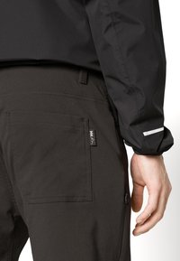 Crna vodootporna jakna glatke teksture, sa vidljivom oznakom loga i kontrastnim bijelim detaljem na rukavu, uparena s tamnim hlačama.