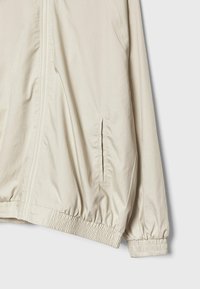 Veste légère beige avec fermeture éclair frontale, poignets et ourlet élastiques, et poches latérales inclinées sur fond uni.