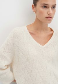 Frau trägt einen weißen, strukturierten V-Ausschnitt-Pullover mit silbernen Creolen und steht vor einer schlichten weißen Wand.
