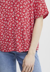 Rote Bluse mit floralen Mustern aus einem strukturierten, gewellten Stoff. Kurze Ärmel, lockere Passform, mit kleinen weißen und schwarzen Blumenmustern. Kombiniert mit blauen Jeans.