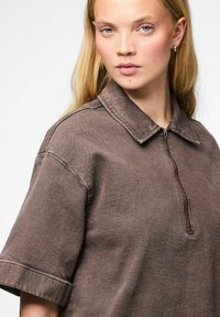 Chemise brune à fermeture éclair avec col, manches courtes et tissu texturé. Présente des coutures visibles et une coupe décontractée.