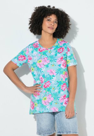 Kvinde med krøllet hår iført en blå t-shirt med blomsterprint og lyse denimbukser, står med den ene hånd på hoften mod en ensfarvet baggrund.