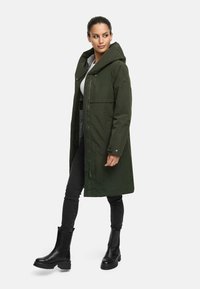 Manteau long vert olive avec une capuche, doté d'une fermeture éclair intégrale, d'accents en bouton et d'un design élégant. Porté par-dessus un haut gris et un jean skinny noir.