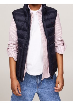 Granatowa kamizelka puchowa o pikowanej fakturze, noszona na jasnej, paskowanej koszuli na guziki oraz białym t-shircie, w zestawie z niebieskimi dżinsami.