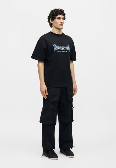 Νέος άνδρας που φοράει ένα μαύρο oversized μπλουζάκι με γράφημα και μαύρο παντελόνι cargo με αθλητικά παπούτσια, σε λευκό φόντο.