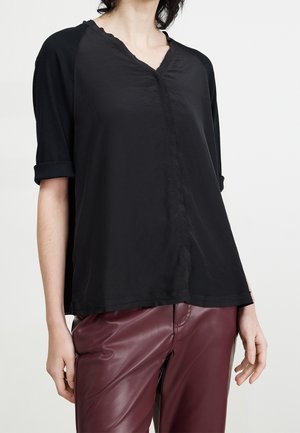Woman wearing a loose black V-neck top with rolled sleeves paired with high-waisted maroon leather pants, standing against a plain background.