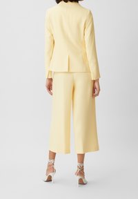Blazer ajusté jaune et pantalons à jambes larges, tissu lisse, design structuré, détail d'emplacement à l'arrière, associé à des talons hauts blancs à brides.