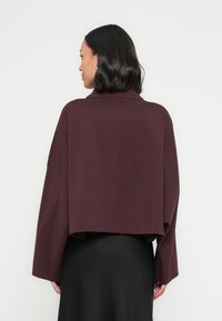 Bordowa krótka kurtka z wysokim kołnierzem, szerokimi rękawami i gładką teksturą, zestawiona z czarną spódnicą. Czysty design bez wzorów.