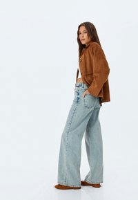 Chaqueta corta de ante marrón con cuello redondeado, combinada con jeans de pierna ancha azul claro y calzado marrón. Diseño sencillo y ajuste relajado.