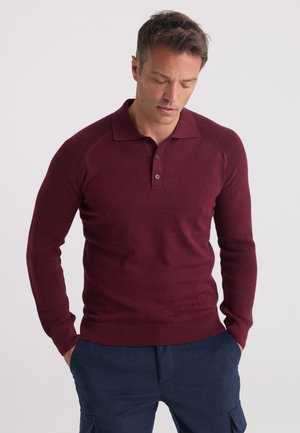 Man wearing a long-sleeve burgundy polo sweater and dark blue pants, looking downward with hands in pockets against plain background.