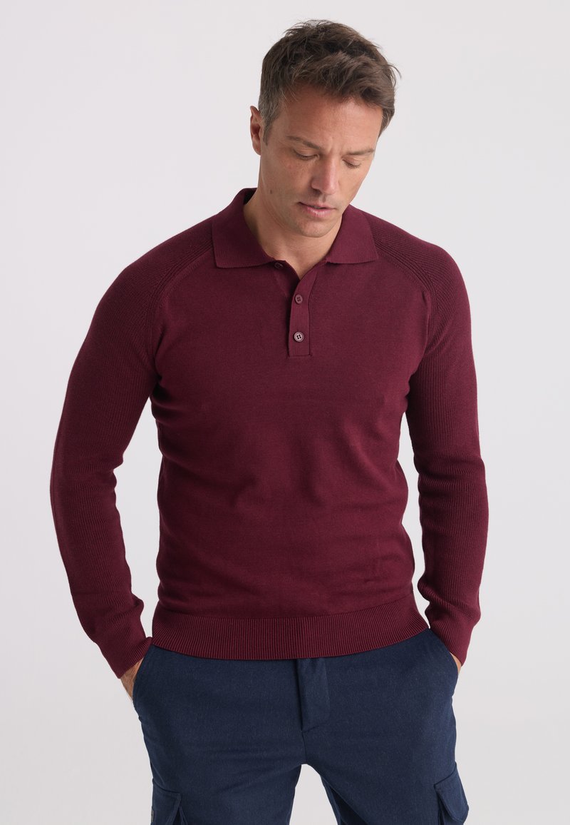 Homme portant un pull polo bordeaux à manches longues et un pantalon bleu foncé, regardant vers le bas avec les mains dans les poches, sur un fond uni.