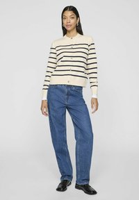 Bežs adīts kardigans ar tumši ziliem svītrām, apaļo kakla izgriezumu un pogu aizdari, kopā ar brīviem ziliem džinsiem un melniem apaviem.