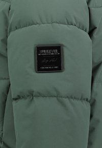 Grüne Pufferjacke aus strukturiertem Material mit gesteppten Abschnitten und einem schwarzen Logo-Patch am Arm. Glattes, modernes Design.