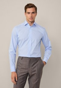 Lichtblauwe blouse met lange mouwen en dunne witte strepen. Heeft een klassieke kraag en een knoopsluiting aan de voorkant, gecombineerd met gemusterde bruine broek.