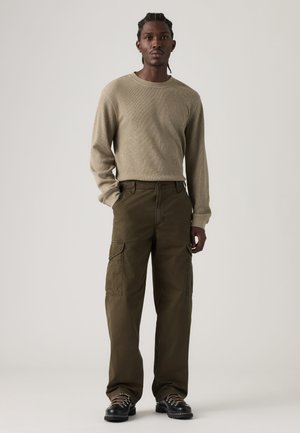 Mann mit geflochtenem Haar steht, trägt einen beigen Langarmpullover, olivfarbene Cargohosen und schwarze Wanderschuhe vor einem einfarbigen hellen Hintergrund.