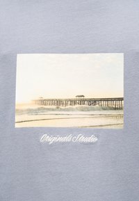 Γαλάζιο μπλουζάκι με ορθογώνιο ανοιχτόχρωμο γραφικό μιας αποβάθρας δίπλα στο νερό, με διακριτικά κύματα και το κείμενο "Originals Studio" σε λευκό κάτω από αυτό.