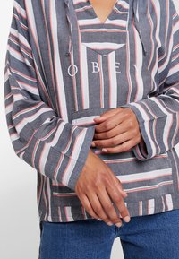 Grau gestreifter Pullover mit rosa und weißen Akzenten, V-Ausschnitt, langen Ärmeln und einer Vordertasche, auf der das Wort "OBEY" in Weiß steht.