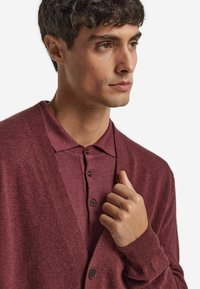 Burgunderfarbener Cardigan mit V-Ausschnitt, Knopfverschluss und gerippten Bündchen, über einem passenden Poloshirt mit Kragen und zwei Knöpfen getragen.