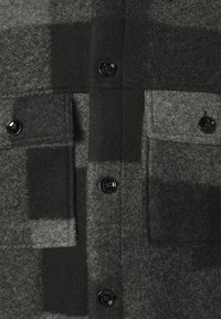 Tela de abrigo de lana negra y gris con un gran patrón de cuadros, dos bolsillos de solapa con botones y una fila de botones negros brillantes en el centro.