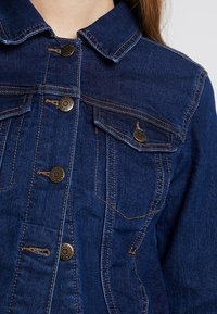 Mörkblå denimjacka med krage, framfickor och metallknappar. Har kontrastsömmar och en strukturerad design.