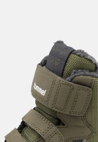 Zapato de invierno color verde oliva con tejido texturizado, correas de velcro a juego, forro de felpa gris y una etiqueta de lengüeta negra con logo dorado y diseño de copo de nieve.
