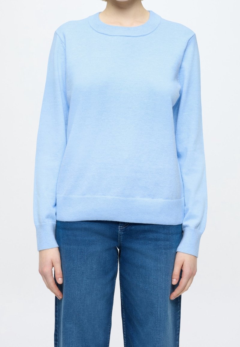 Personne portant un pull bleu clair à manches longues et un jean bleu moyen, debout devant un fond blanc uni.