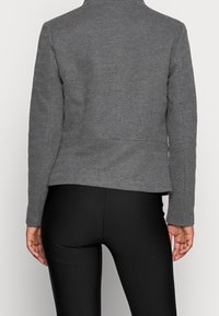 Donna che indossa un blazer grigio a spina di pesce aderente e leggings neri, mostrata di spalle su uno sfondo chiaro e semplice.